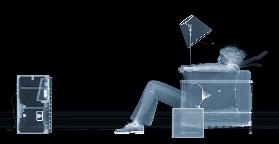 A limited edition Chromaluxe print by Nick Veasey of an X-Rayed person sat in a chair with a lamp and drink next to them all being blown over, the title is Blown Away