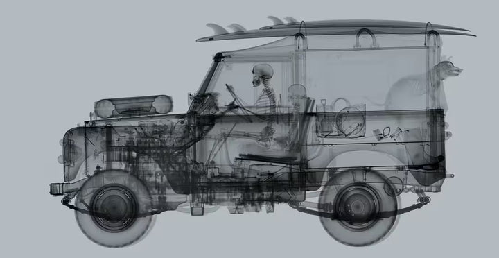 A limited edition Chromaluxe print by Nick Veasey of an X-Rayed vehicle with a skeleton person sat in the front and a skeleton dog leaning out of the back of the car along with beach items scattered around the car, the title is Land Rover Surfer- Mid Grey
