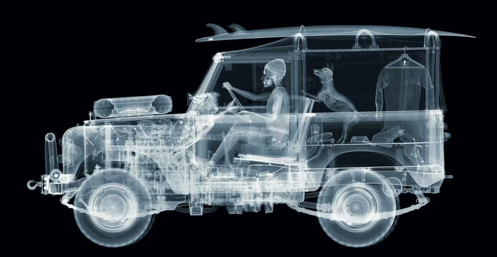 A limited edition Chromaluxe print by Nick Veasey of an X-Rayed vehicle with a skeleton person sat in the front and a skeleton dog leaning out of the back of the car along with beach items scattered around the car, the title is Land Rover- Surfs Up