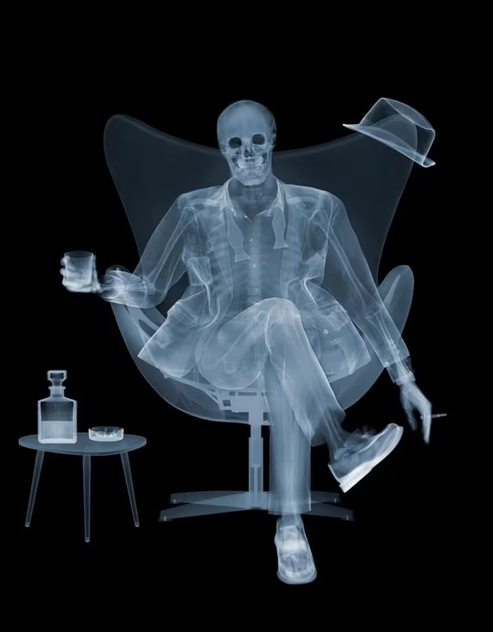 A limited edition Chromaluxe print by Nick Veasey of an X-Rayed ratpack member sat on a big chair with a drink in their hand and a table with a drink. next to them, they are also holding a cigarette, the title is Rat Pack III