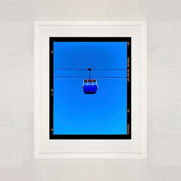A white framed limited edition photograph by Richard Heeps using vibrant colours, the title is Summit Cable Car (Blue)