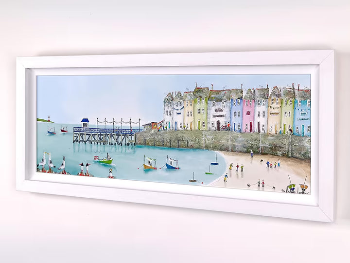 A Rebecca Lardner limited edition piece of a row of colourful houses with a pier at the end and people on the beach and boats on the sea with puffins, seagulls, and avocets on the rocks, the title is Tide and Seek