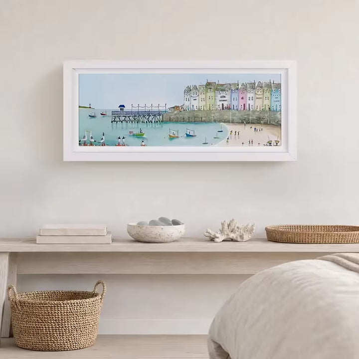 A Rebecca Lardner limited edition piece of a row of colourful houses with a pier at the end and people on the beach and boats on the sea with puffins, seagulls, and avocets on the rocks, the title is Tide and Seek